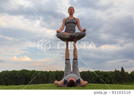 Man lying on grass and balancing woman in his feet. Young couple doing acro yoga in park Man lying on grass and balancing woman in his feet. Young couple doing acro yoga in park 93103510