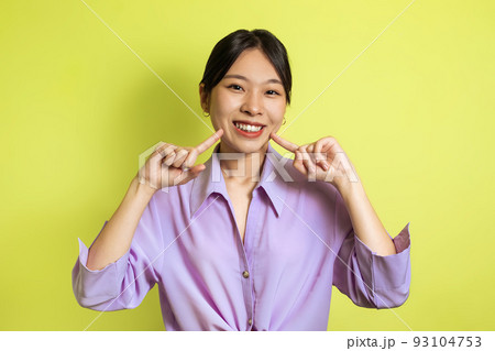 Toothy Asian Female Pointing Fingers At Her Smile, Yellow Background Toothy Asian Female Pointing Fingers At Her Smile, Yellow Background 93104753