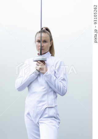 Caucasian woman in white fencing costume hold epee in front of face, wears jacket, pants. Sports, healthy lifestyle, professional coach. Vertical plane. 93105262