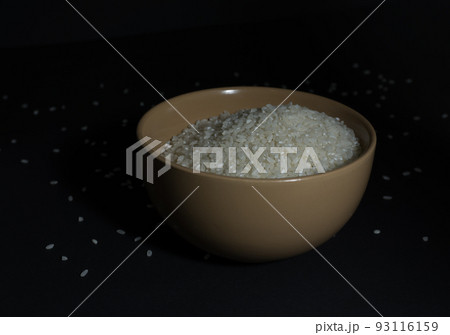 White uncooked rice in a bowl with scattered grains nearby on a black background White uncooked rice in a bowl with scattered grains nearby on a black background 93116159