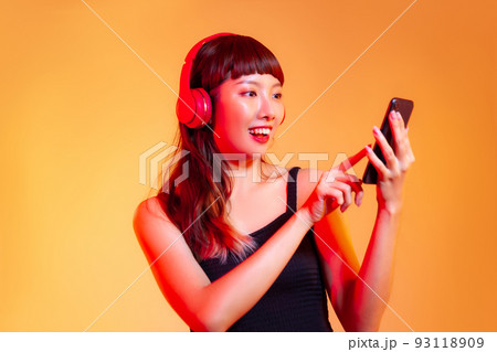 Young asian woman in black tank top wearing red headphones and holding smartphone chatting and shopping online on the orange background 93118909