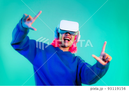 Young asian girl pink color hairstyle in blue sweatshirt wearing white vr headset finger pointing playing cheering and touching on the green screen background. Cyborg and Metaverse concept 93119156