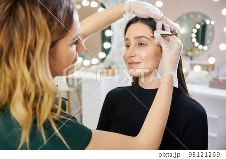 Smiling young woman having eyebrow correction procedure in beauty salon. Female esthetician in sterile gloves shaping woman eyebrows with cosmetic tweezers. 93121269