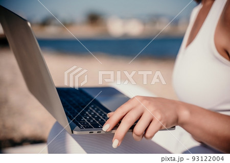 young beautiful woman with dark hair in white overalls sits on the sand near the sea and uses a laptop 93122004