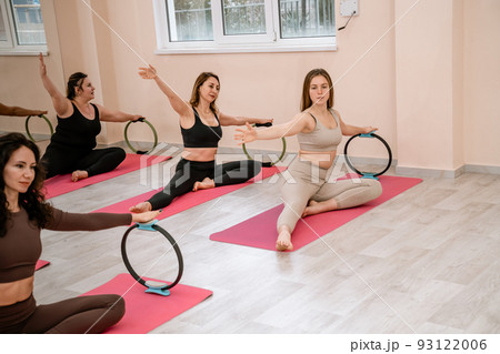 A group of six athletic women doing pilates or yoga on pink mats in front of a window in a beige loft studio interior. Teamwork, good mood and healthy lifestyle concept. 93122006