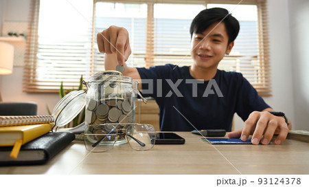 Smiling young asian male putting coin into the jar. Save money for future, retirement and financial planning concept 93124378