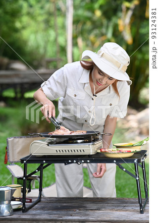 Young woman wearing casual clothing and hat grilled bbq near the river bank surrounded by beautiful nature 93124381