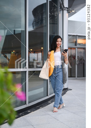 Full length portrait of young woman with shopping bags using smart phone and walking in the city street at the evening 93124382