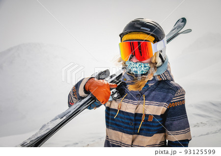 Portrait of a pretty and active woman skier, wearing a mask and holding skis in her hands, active winter holidays 93125599