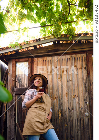 Pretty woman, eco farmer, winemaker, winegrower wearing a straw hat and a beige apron standing by a wooden countryside house and relaxing with a glass of red wine after harvesting grapes on vineyards 93135402