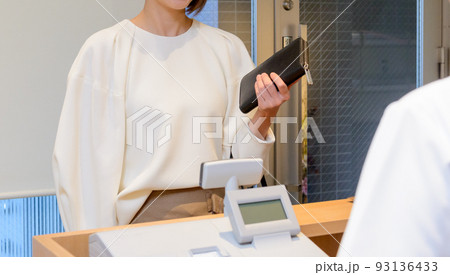 財布を手に持ったマスクを着けた女性が、アクリル板が設置された受付で会計するシーン 93136433