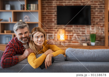 Happy adult european man and woman looking at camera, enjoy relaxing in living room interior with tv with blank screen Happy adult european man and woman looking at camera, enjoy relaxing in living room interior with tv with blank screen 93138678