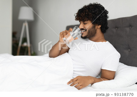 Happy young indian man holding glass of water in bed Happy young indian man holding glass of water in bed 93138770