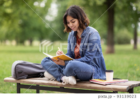 Arab female student writing in notebook while sitting on bench in park 93139023