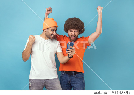 Portrait of two cheerful extremely happy young adult hipster men clenched fists while using mobile phone, screaming yes, betting and winning. Indoor studio shot isolated on blue background. Portrait of two cheerful extremely happy young adult hipster men clenched fists while using mobile phone, screaming yes, betting and winning. Indoor studio shot isolated on blue background. 93142397