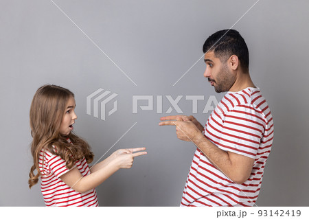 Portrait of surprised astonished father and daughter in striped T-shirts standing pointing at each other, looking with big eyes, choosing turn. Indoor studio shot isolated on gray background. Portrait of surprised astonished father and daughter in striped T-shirts standing pointing at each other, looking with big eyes, choosing turn. Indoor studio shot isolated on gray background. 93142419