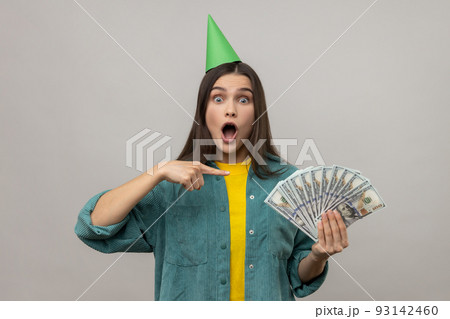 Amazed excited rich woman in party cone on head pointing at money, looking surprised by lottery win, celebrating big profit, wearing casual style jacket. Indoor studio shot isolated on gray background Amazed excited rich woman in party cone on head pointing at money, looking surprised by lottery win, celebrating big profit, wearing casual style jacket. Indoor studio shot isolated on gray background 93142460