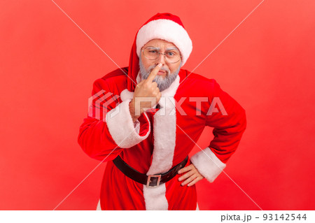 Elderly man with gray beard wearing santa claus costume touching nose, gesturing you are liar, being distrustful of talk, suspecting falsehood. Indoor studio shot isolated on red background. 93142544