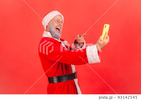 Portrait of elderly man with gray beard wearing santa claus costume standing with smart phone pointing to display, looking with toothy smile. Indoor studio shot isolated on red background. 93142545