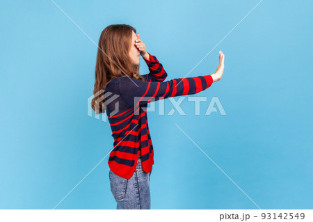 Side view portrait of woman wearing striped casual style sweater standing and afraid, covering her eyes with hand and trying to blocking Indoor studio shot isolated on blue background. Side view portrait of woman wearing striped casual style sweater standing and afraid, covering her eyes with hand and trying to blocking Indoor studio shot isolated on blue background. 93142549