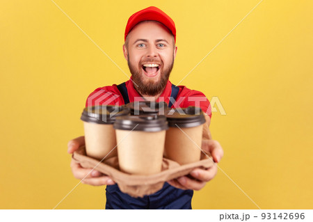 Delivery service. Friendly happy courier man in blue overalls offering coffee, giving drinks in disposable cups, looking at camera with excited look. Indoor studio shot isolated on yellow background. 93142696