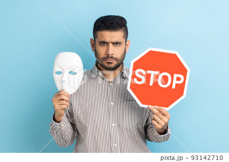 Portrait of serious businessman holding white mask with unknown face and red traffic sign, looking at camera, wearing striped shirt. Indoor studio shot isolated on blue background. Portrait of serious businessman holding white mask with unknown face and red traffic sign, looking at camera, wearing striped shirt. Indoor studio shot isolated on blue background. 93142710
