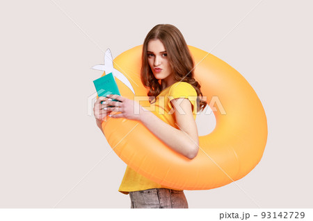 Portrait of tourist teenager girl in yellow T-shirt holding rubber ring, passport document and airplane mockup, looking at camera with pouts lips. Indoor studio shot isolated on gray background. Portrait of tourist teenager girl in yellow T-shirt holding rubber ring, passport document and airplane mockup, looking at camera with pouts lips. Indoor studio shot isolated on gray background. 93142729