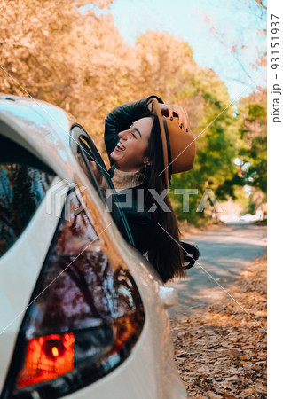 Beautiful young woman carrying hat and looking happy while enjoying road trip. Young woman with long hair in brown hat and sweater looking out from white car holding hat and scarf against autumn 93151937
