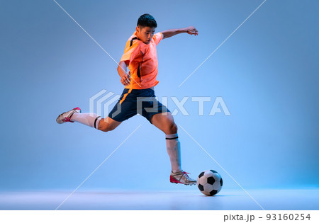 Dynamic portrait of young man, professional football player in motion, training, dribbling ball isolated over blue studio background in neon light 93160254