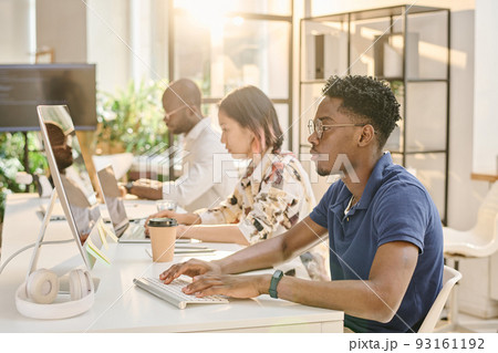 African young programmer in eyeglasses concentrating on his work on computer sitting at table with his colleagues at office 93161192