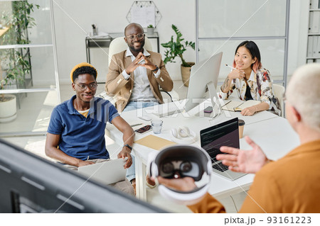 Multiethnic group of colleagues sitting at table at meeting and listening to speaker telling about new app of virtual reality glasses 93161223