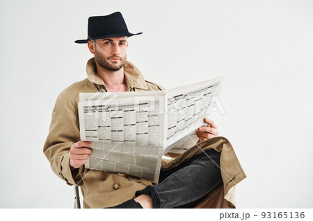 Young handsome man in a trench coat and hat reading newspaper sitting on chair over white background 93165136