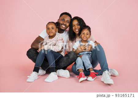 Portrait of smiling african american family of four sitting on floor over pink studio background 93165171