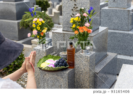 お墓参りに来てきれいなお花とお供え物をして手を合わせている中年女性 お墓参りに来てきれいなお花とお供え物をして手を合わせている中年女性 93165406