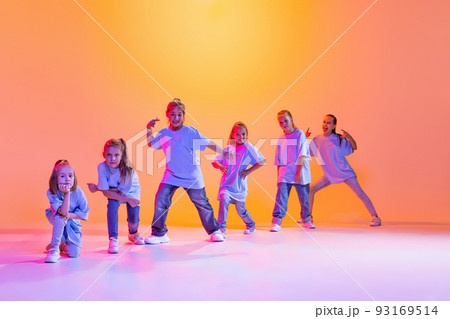 Dance group of happy, active little girls in t-shirts and jeans in action isolated on orange background in neon. Concept of music, fashion, art, childhood Dance group of happy, active little girls in t-shirts and jeans in action isolated on orange background in neon. Concept of music, fashion, art, childhood 93169514