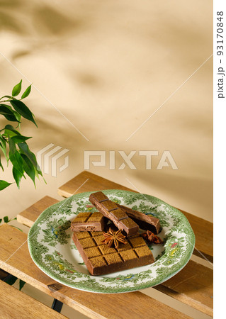a broken chocolate bar with spices on a vintage plate on a wooden table on a sunny day. Beige background 93170848