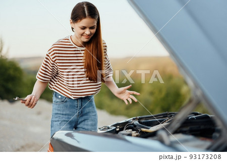 A woman traveler stands by her old car with the hood open and repairs the car engine with a wrench, unscrewing car parts A woman traveler stands by her old car with the hood open and repairs the car engine with a wrench, unscrewing car parts 93173208