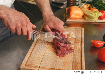 Close up of chef cutting fresh raw meat on wooden board in modern kitchen of restaurant Close up of chef cutting fresh raw meat on wooden board in modern kitchen of restaurant 93174091