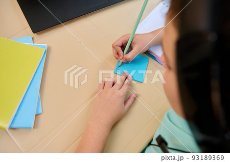 Overhead view of a schoolchild during a video conference with the teacher in virtual class, with selective focus on his hands holding a pencil and making notes during an online lesson on the laptop 93189369