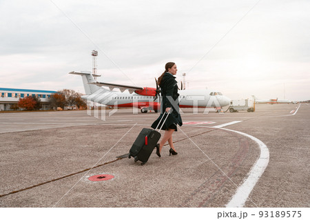 Woman with travel suitcase walking down airfield on background of airplane Woman with travel suitcase walking down airfield on background of airplane 93189575