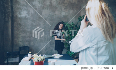 Attractive girl is posing with large green plant while female colleague photogrpahing her on digital camera in modern lof office. Women are having fun and laughing during coffee break 93190813