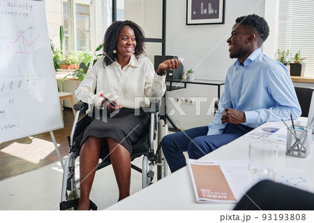 African businesswoman sitting on wheelchair near the flipchart and discussing business strategy with her partner at office African businesswoman sitting on wheelchair near the flipchart and discussing business strategy with her partner at office 93193808