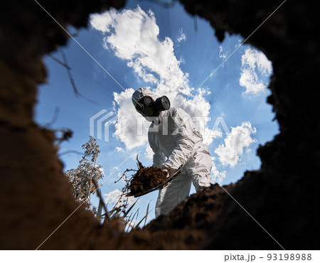 View from inside pit of ecologist digging pit by shovel and planting tree, wearing gas mask and protective overalls outdoors, on background of blue cloudy sky. 93198988