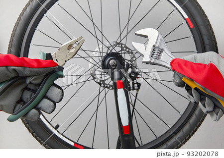 Hands in protective gloves with adjustable wrenches and pliers on the background of a bicycle wheel. Bicycle repair at home 93202078