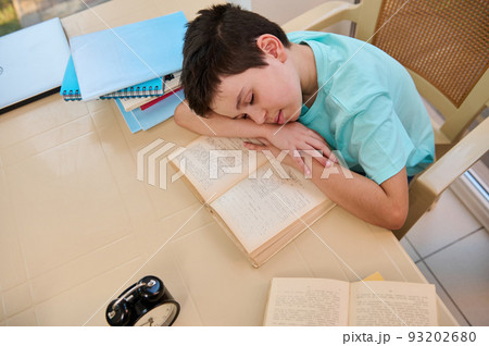 Top view of a tired sleepy schoolboy falling asleep on a textbook while doing homework. Homeschooling. Daily routine of a student. Learning disability. Difficulties in restoring the school regime Top view of a tired sleepy schoolboy falling asleep on a textbook while doing homework. Homeschooling. Daily routine of a student. Learning disability. Difficulties in restoring the school regime 93202680