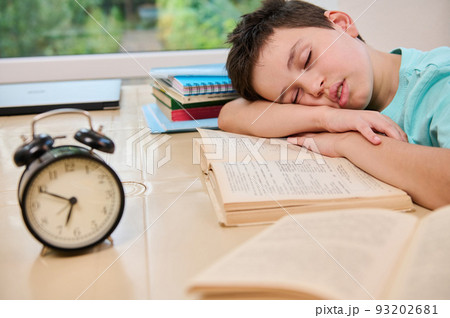 Blurred black alarm clock on the background of a tired sleepy schoolboy falling asleep on a textbook while doing homework. Teenage boy having learning disability. Back to school 93202681