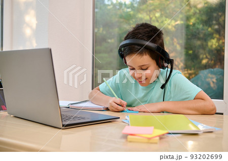 Smart happy multi-ethnic kid, schoolboy in headphones, makes notes while talking with remote web teacher on social distance video conference call elearning class on computer. Children learning at home 93202699