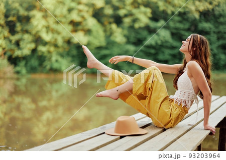 A young hippie woman sits on a lake bridge wearing stylish eco clothes and smiling A young hippie woman sits on a lake bridge wearing stylish eco clothes and smiling 93203964