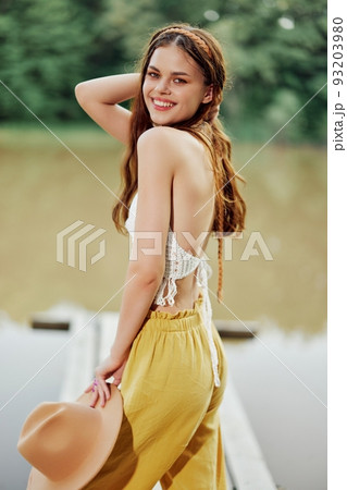 A young woman smiling in an image of a hippie and eco-dress dancing in nature by the lake wearing a hat and yellow pants in the summer sunset 93203980