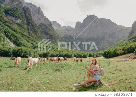 Young woman sitting and relaxing on alpine mountain looking the cow eating a grass in the summer. Travel, Lifestyle Concept. Beautiful woman enjoys views of the alpine village in the Alps mountains Young woman sitting and relaxing on alpine mountain looking the cow eating a grass in the summer. Travel, Lifestyle Concept. Beautiful woman enjoys views of the alpine village in the Alps mountains 93205971